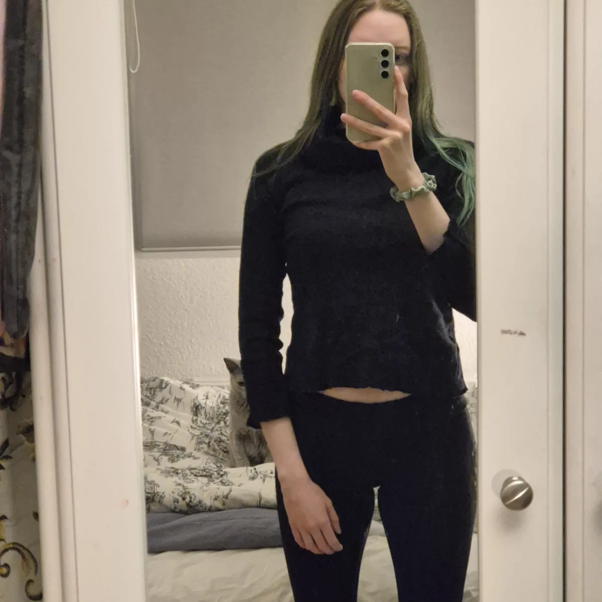 A person with long hair, partially dyed green, takes a mirror selfie in a bedroom while wearing a black cropped turtleneck…
