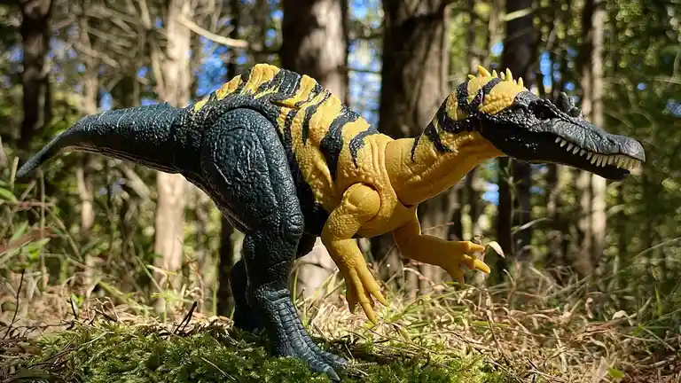 A yellow and black striped plastic dinosaur toy, resembling a raptor, is posed in a forest setting on mossy ground. The toy…