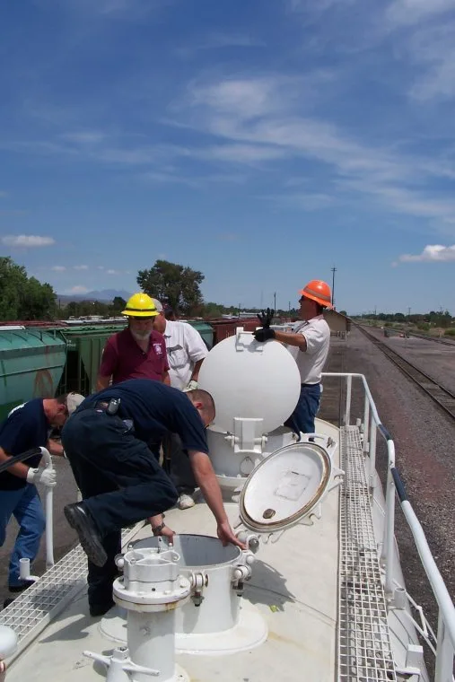 Five men in work clothes and hard hats are on top of a large white industrial tank under a blue sky. One man is bent over…
