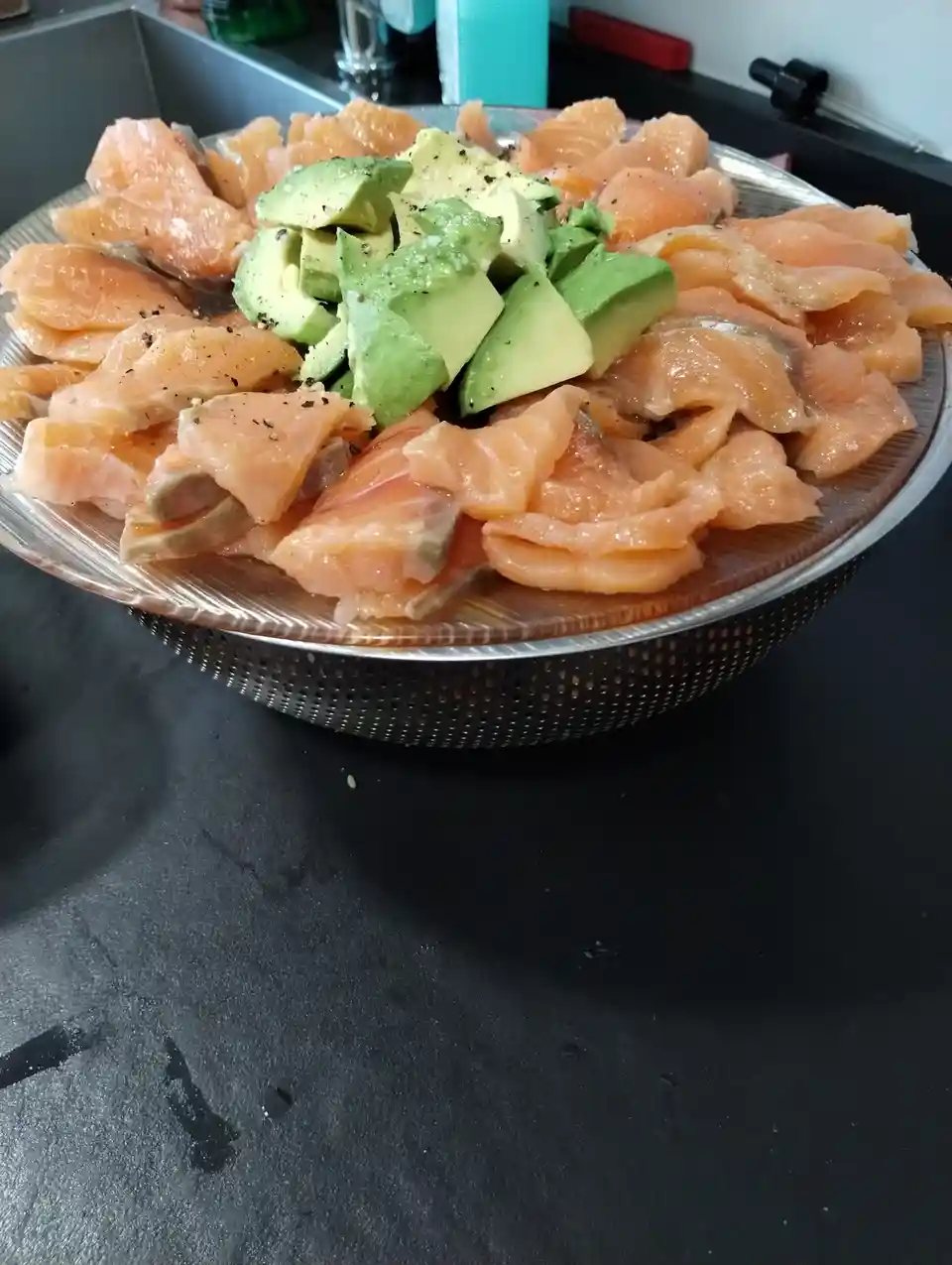 A metal colander filled with raw salmon slices and chopped avocado, seasoned with black pepper, sits on a dark kitchen…