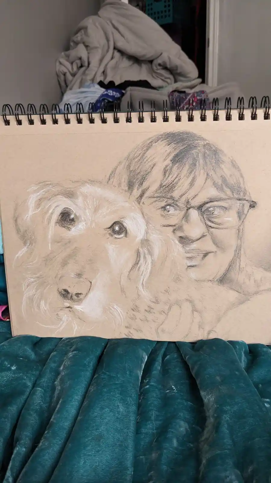 A pencil sketch in a spiral-bound sketchbook shows a detailed portrait of a person with glasses next to a dog, both rendered…