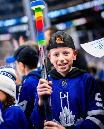 A young boy wearing a blue Toronto Maple Leafs hockey jersey and a black baseball cap smiles at the camera while holding a…