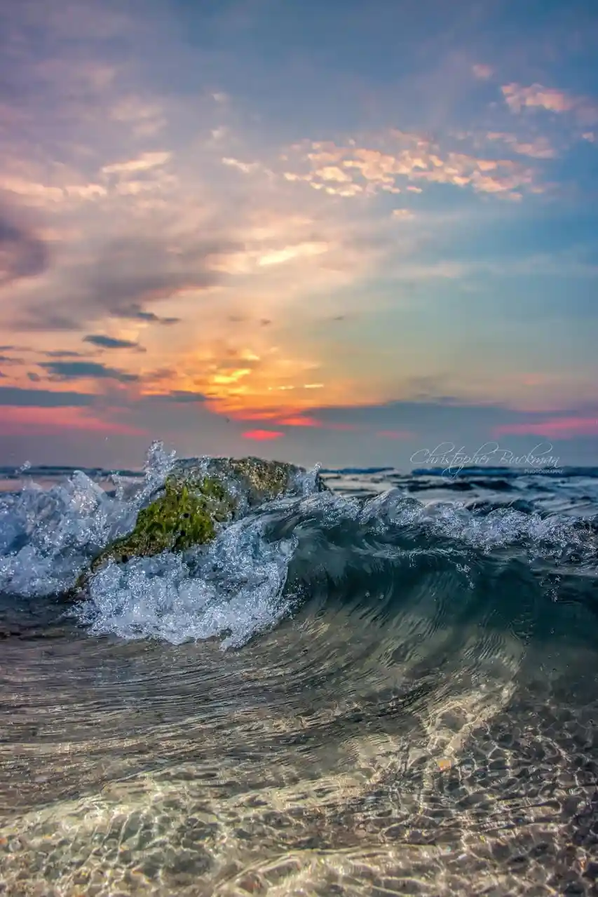 A serene sunset photograph shows ocean waves crashing over a moss-covered rock, with vibrant orange, pink, and blue hues in…
