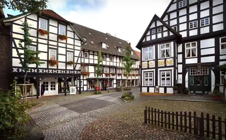 A picturesque cobblestone courtyard surrounded by traditional black-and-white half-timbered German buildings with flower…