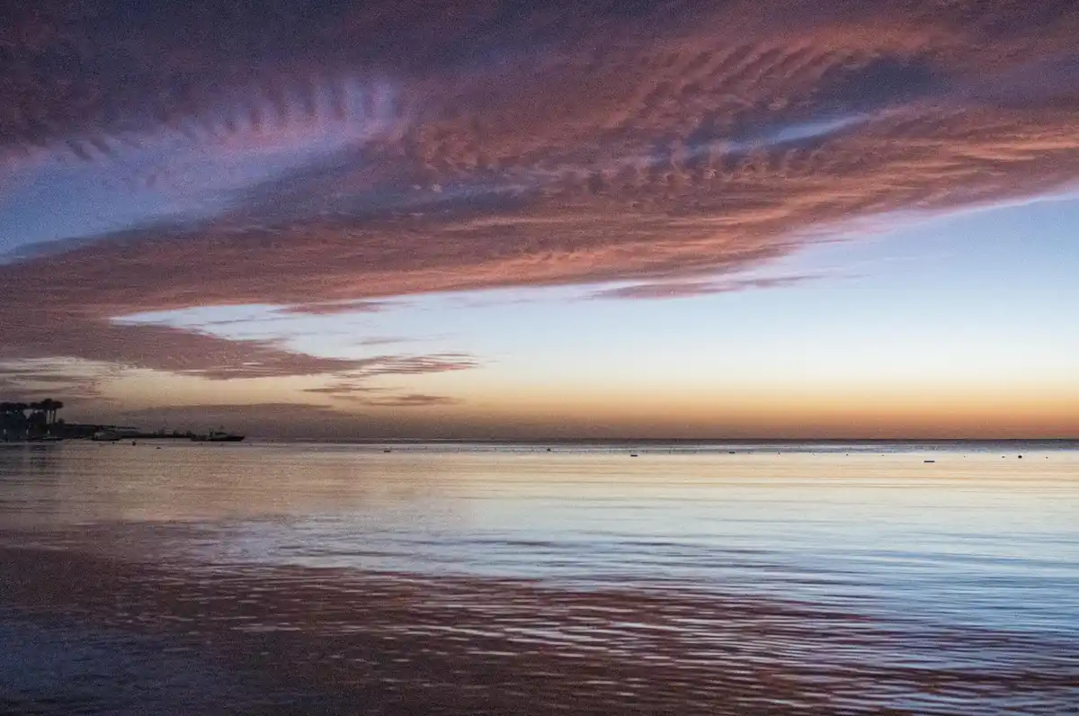 A serene sunset over a calm body of water, with vibrant pink, purple, and orange clouds reflecting on the surface. A distant…