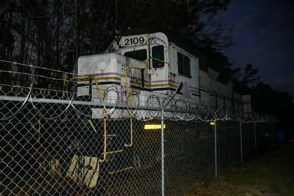 A weathered, white train engine numbered 2109 sits behind a chain-link fence topped with coiled razor wire at dusk. The…