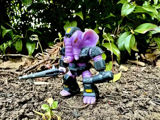 A purple elephant toy with black mechanical armor and a weapon stands in dirt surrounded by green foliage. The toy has a…