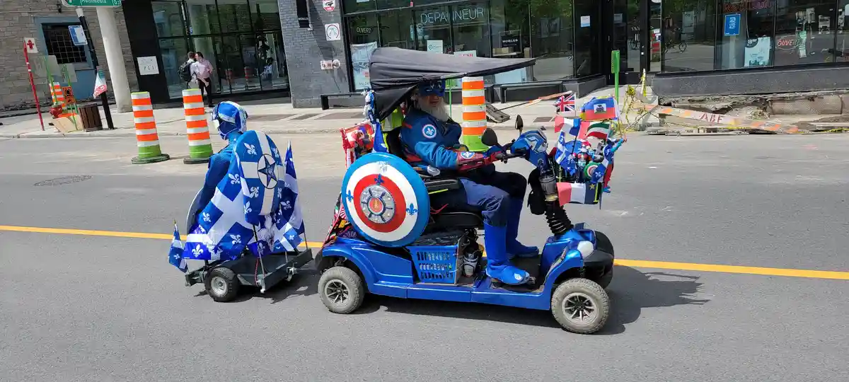 A man in a blue superhero costume with a Captain America-style shield sits in a blue mobility scooter decorated with…