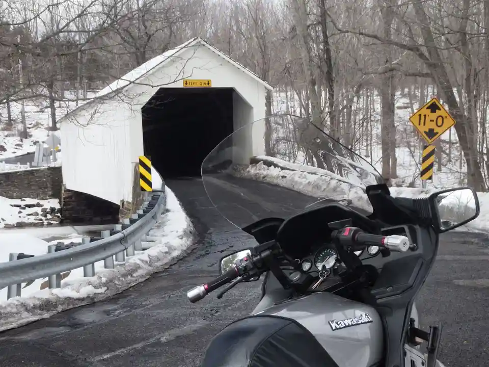 A silver Kawasaki motorcycle is parked on a snowy road facing a white covered bridge. Two yellow signs indicate the bridge's…