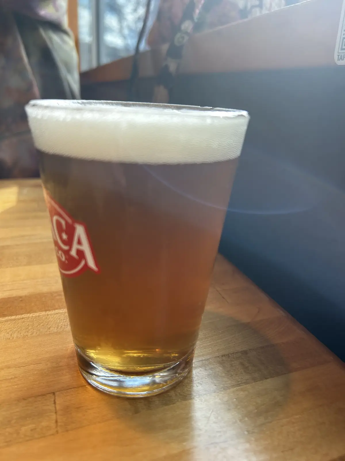 A pint glass filled with amber beer and a thick white head sits on a wooden table, with a red logo partially visible on the…