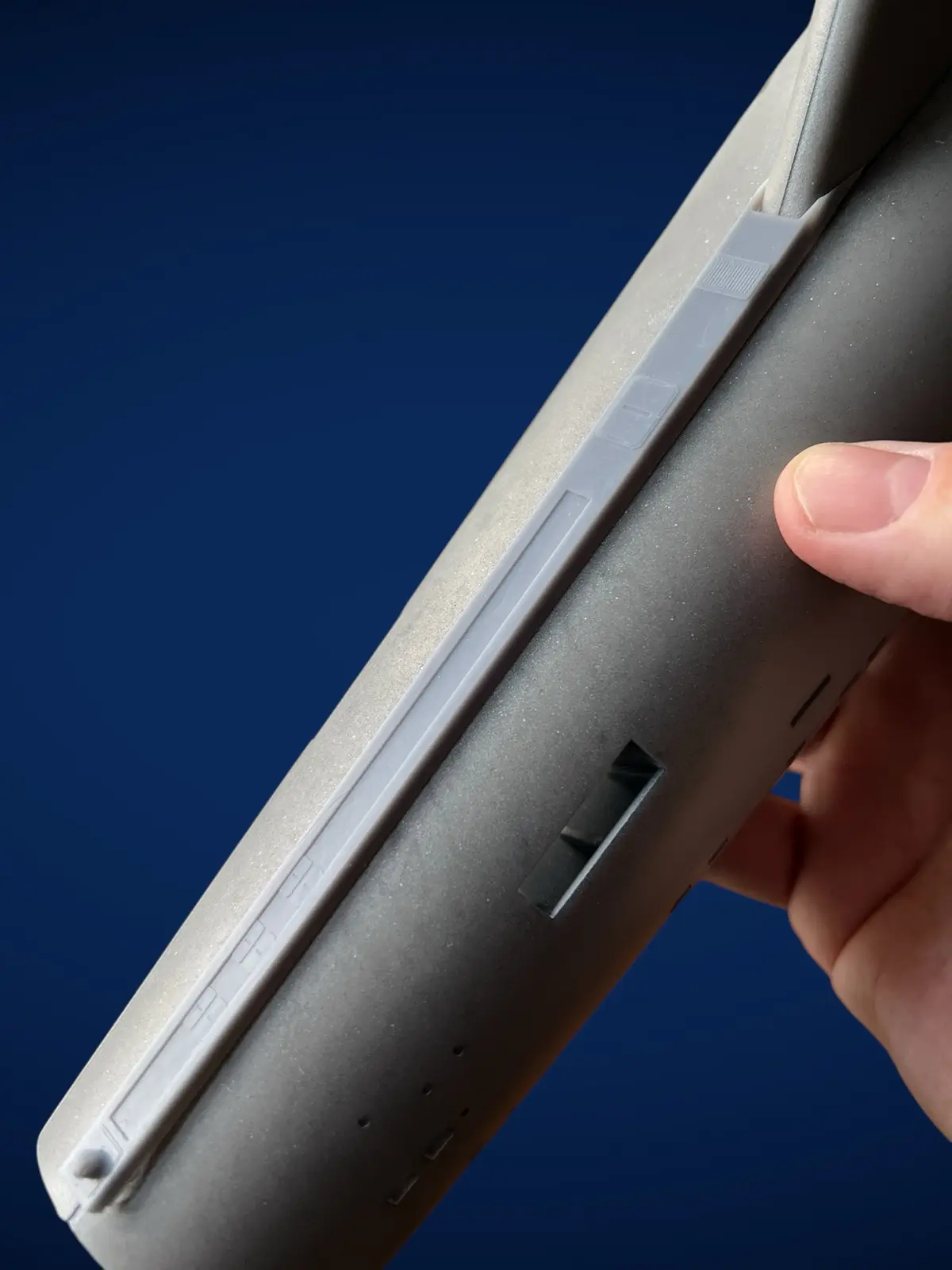 A close-up photo shows a hand holding a gray remote control against a dark blue background, highlighting a small rectangular…