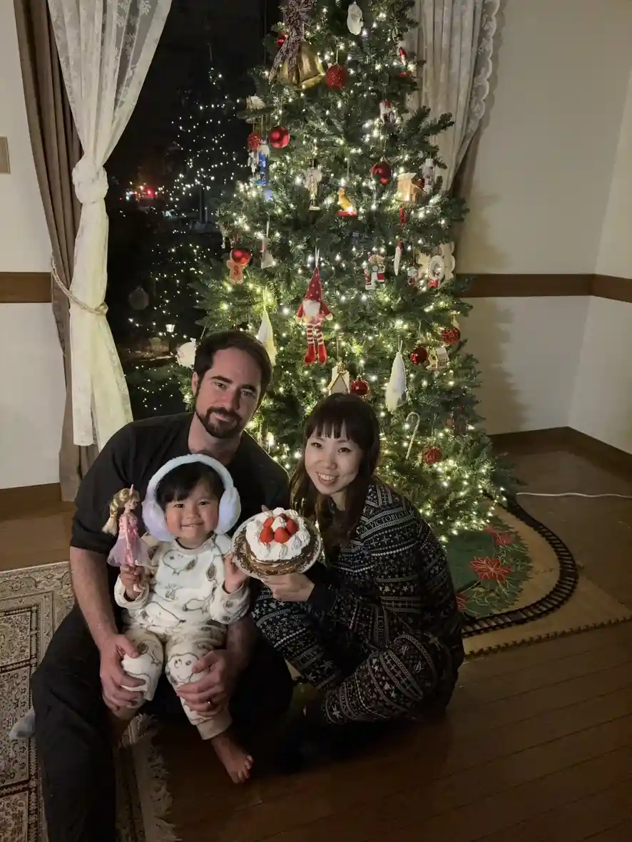 A family of three poses in front of a brightly lit, decorated Christmas tree. The father holds a toddler wearing white…