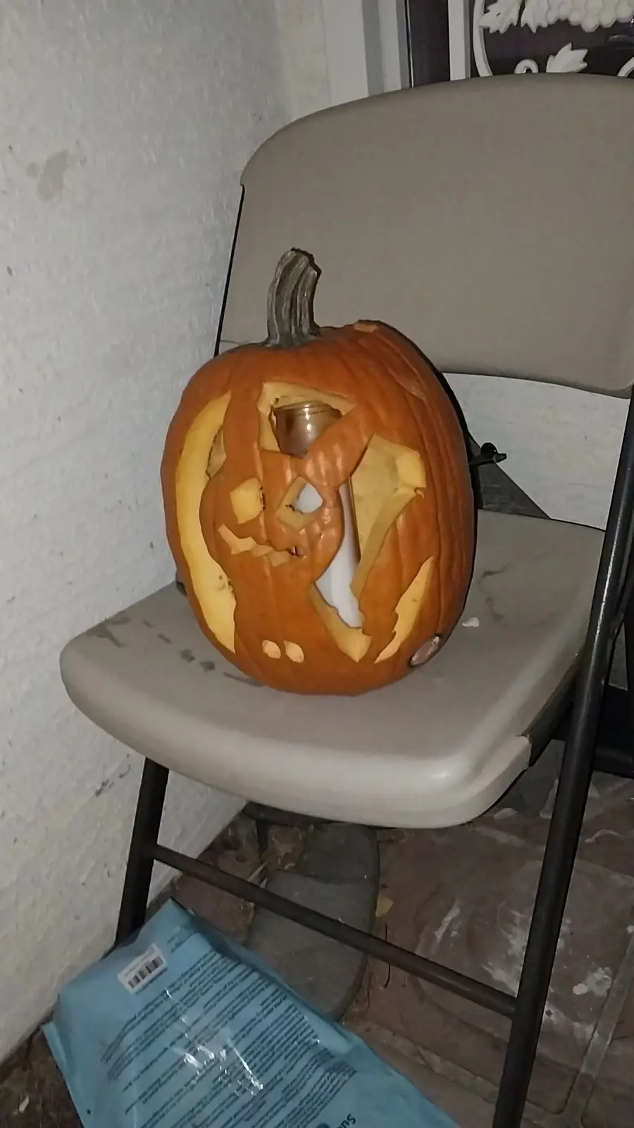 A carved orange pumpkin with a crude, asymmetrical face sits on a beige folding chair. The pumpkin has jagged eye and mouth…