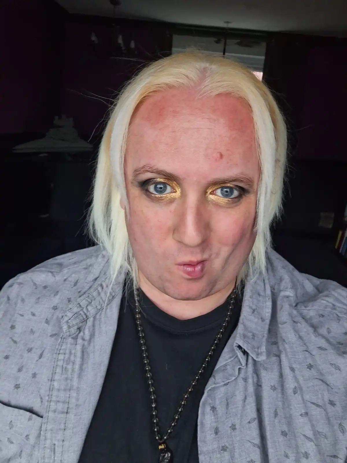 A person with platinum blonde hair and heavy gold eyeshadow stares directly at the camera with a puckered lip expression…