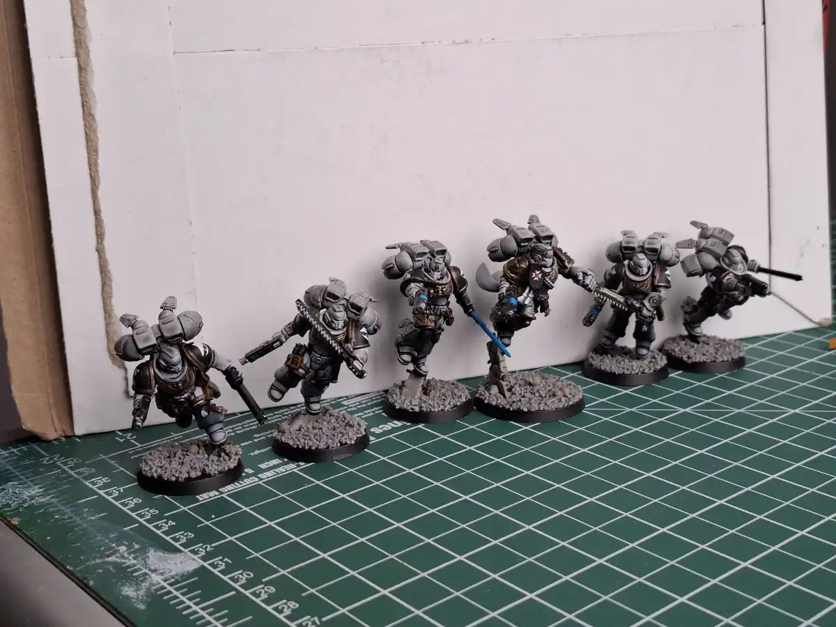 Five painted miniature sci-fi soldiers in grey armor stand in a line on a green grid mat against a white wall. Each figure…