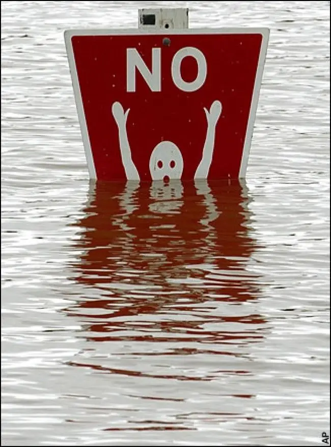 A red, rectangular 'NO' traffic sign is partially submerged in floodwater, with a white ghost-like figure drawn on its…