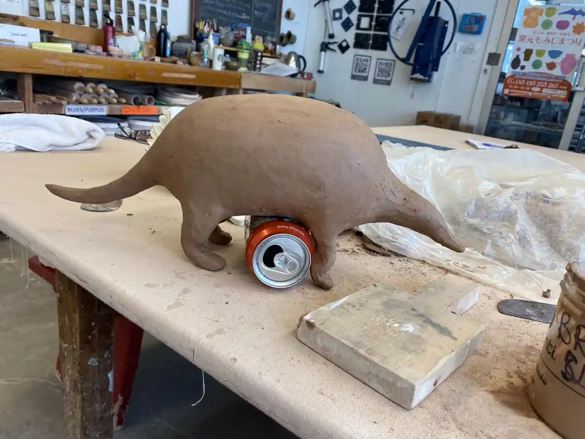 A clay sculpture of a small, four-legged animal with a long snout and tail is positioned on a workbench, with its body…