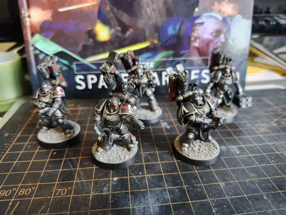 Five painted miniature Space Marines in power armor stand on a grid mat, with a 'Warhammer 40,000: Space Marines' box…