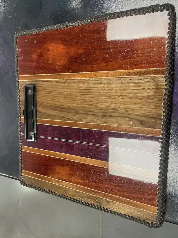 A handmade wooden clipboard with a decorative braided leather border and alternating wood grain stripes in red, tan, and…