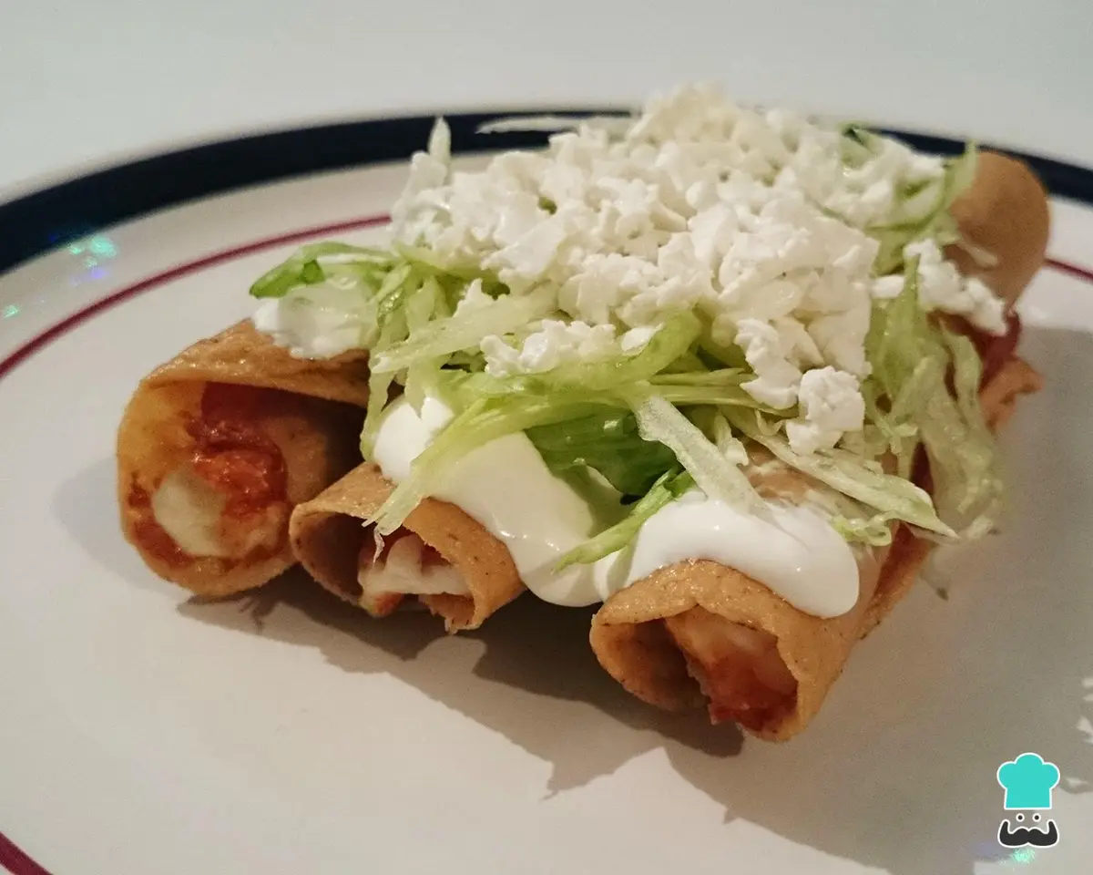 Three fried taquitos on a white plate with a blue and red rim, topped with shredded lettuce, crumbled white cheese, and a…