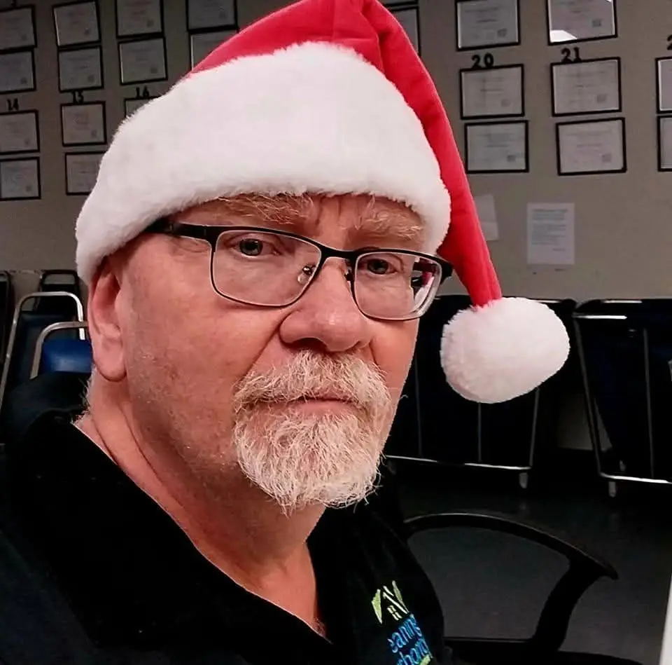 A middle-aged man with glasses and a white beard wears a red and white Santa hat while looking directly at the camera with a…