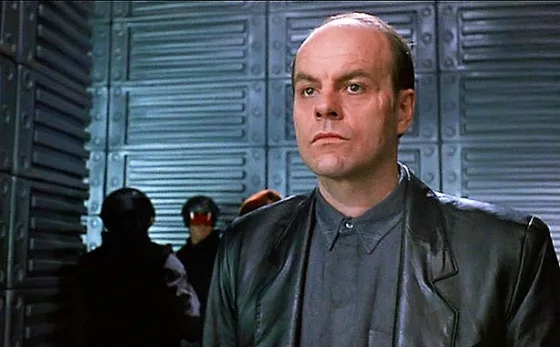 A man with a balding head and intense expression wears a black leather jacket over a gray shirt, standing in front of a…