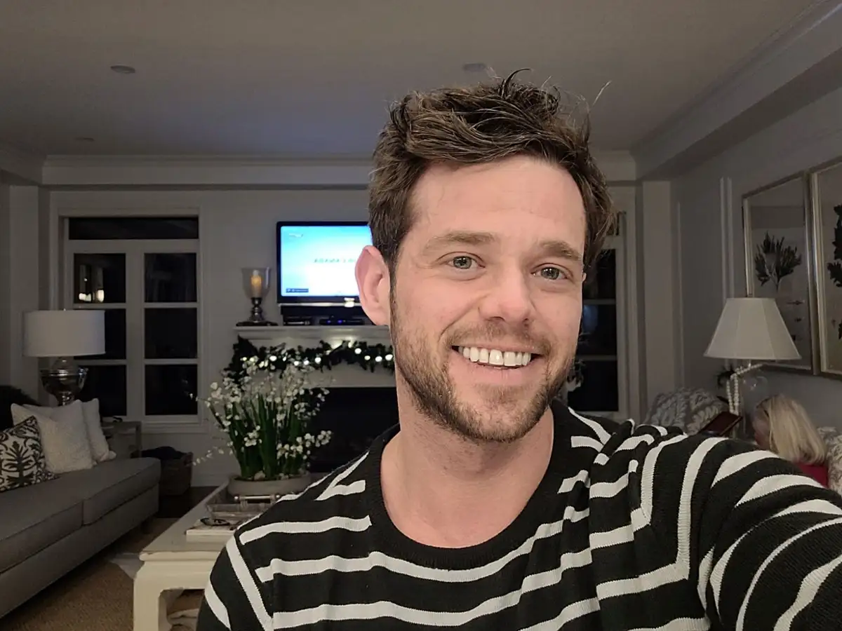 A man with short brown hair and a beard, wearing a black and white striped shirt, smiles broadly at the camera in a selfie…