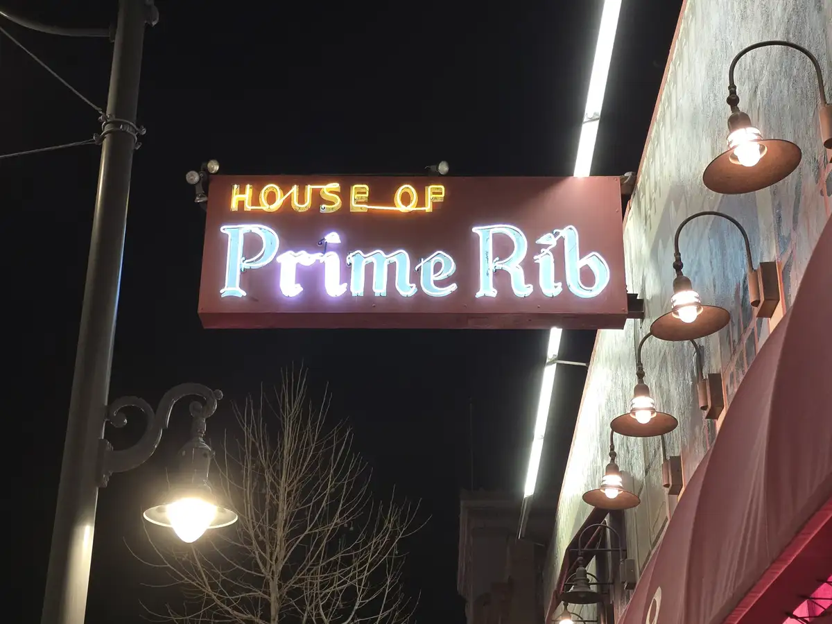 A nighttime photo shows a glowing neon sign for a restaurant called 'HOUSE OF Prime Rib' mounted above a building entrance…