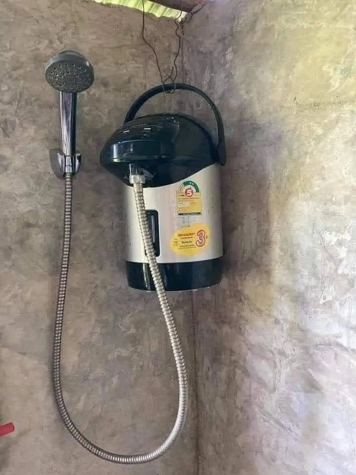 A silver and black Sharp rice cooker is mounted on a tiled wall, with a shower hose and handheld shower head connected to…