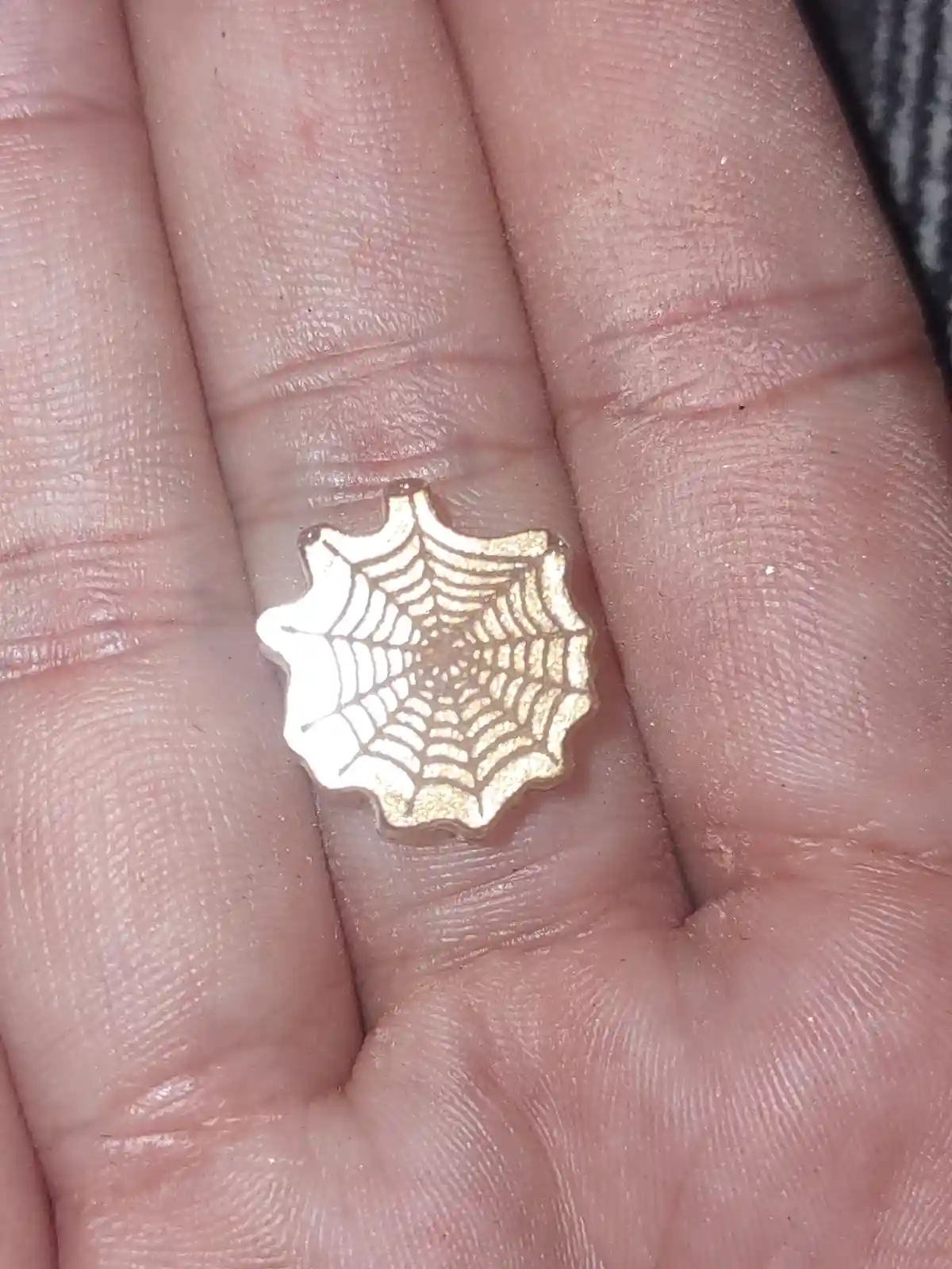 A close-up photo shows a small, gold-colored, spiderweb-shaped pin resting in the center of an open human palm, with visible…