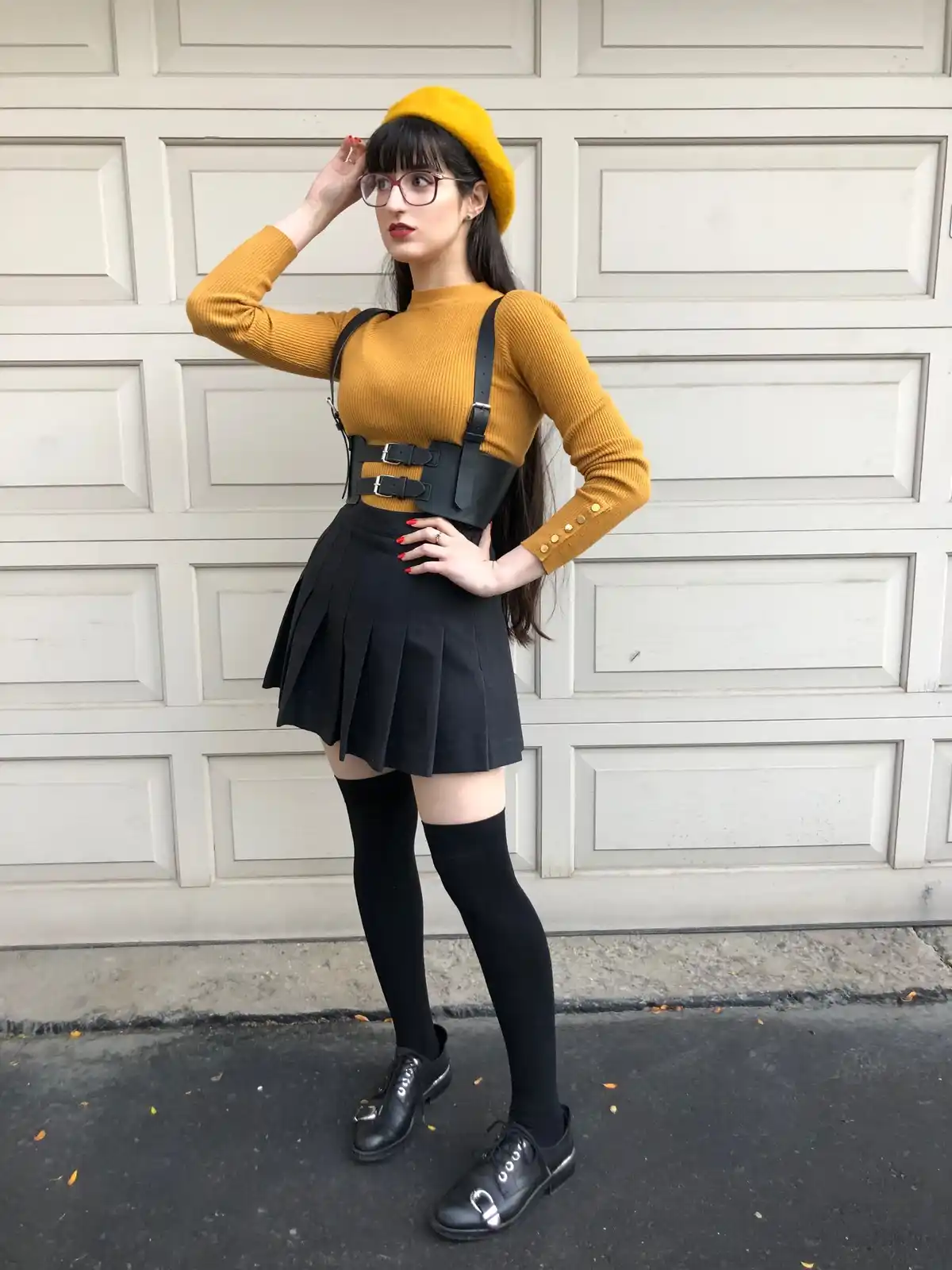 A woman stands in front of a white garage door wearing a mustard yellow sweater, black pleated skirt, black thigh-high…