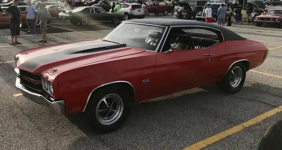 A red classic Chevrolet Chevelle SS muscle car with a black hood and black vinyl roof is parked in a crowded car show lot…