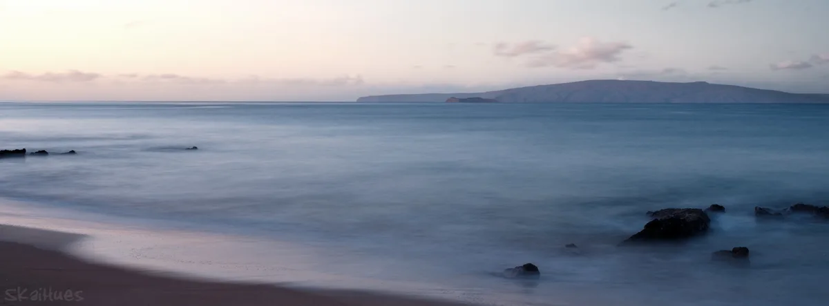 A long-exposure photograph of a calm beach at dusk, showing smooth, misty water flowing around dark rocks, with a distant…