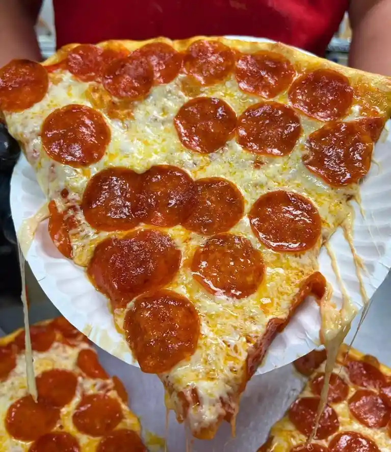 a close-up photo shows a person holding a large, cheesy slice of pepperoni pizza on a paper plate, with melted cheese…