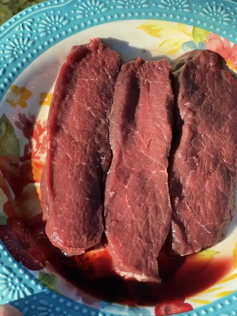 Three raw beef steaks sit on a colorful floral plate with a blue rim, surrounded by pooling red liquid that resembles blood…