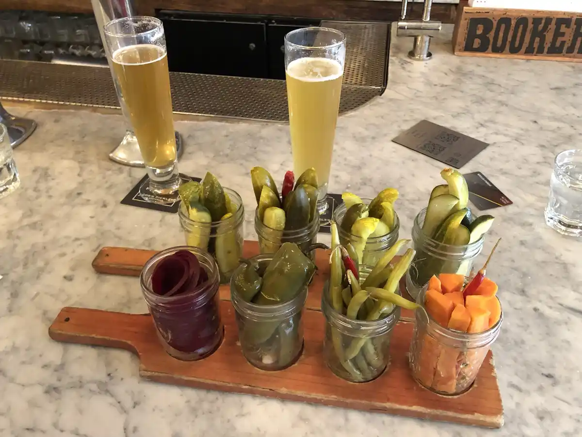 A wooden board holds several small glass jars filled with various pickled foods like pickles, jalapeños, beets, and carrots…
