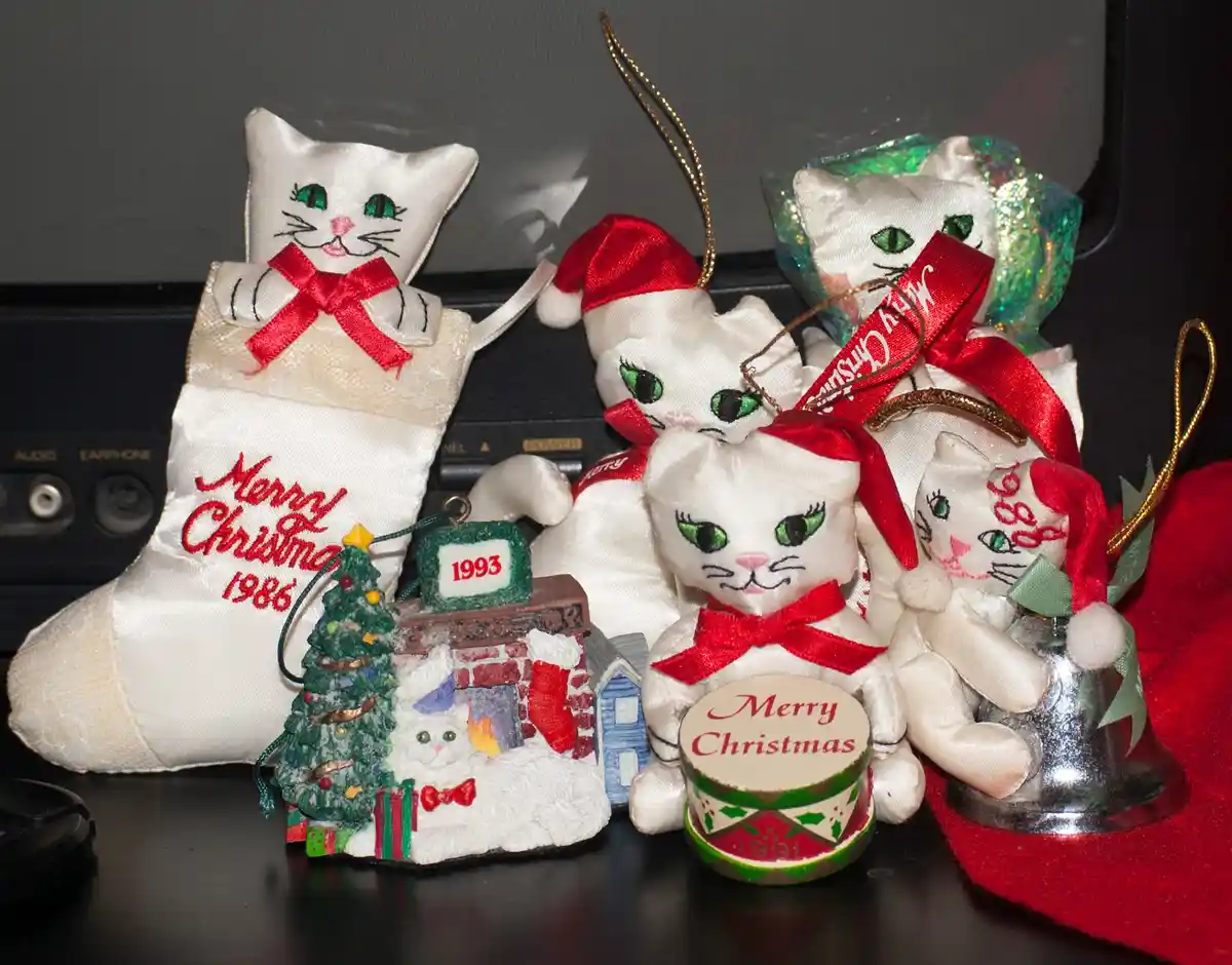 A collection of vintage Christmas ornaments featuring white satin cats with green eyes, red bows, and Santa hats, displayed…