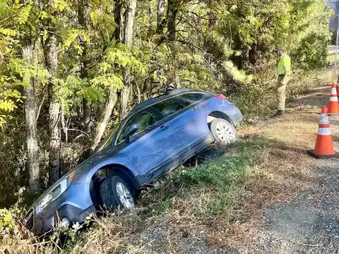 A blue Subaru Outback is partially off-road, tilted into a wooded area with its front end in bushes and rear on gravel. A…