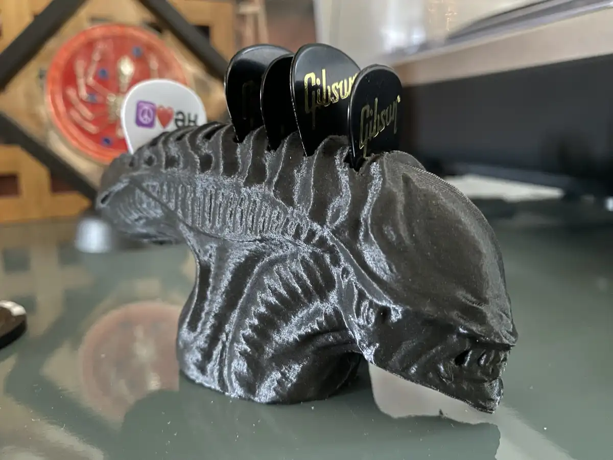 A 3D-printed black Xenomorph head from the movie Alien sits on a glass surface, with four black Gibson guitar picks inserted…