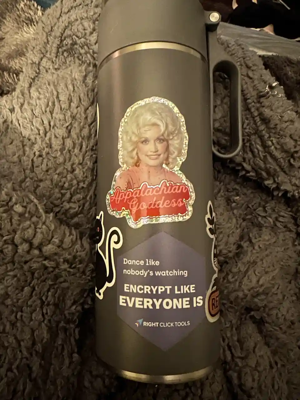 A dark grey insulated water bottle with a handle sits on a fluffy grey blanket. It has a holographic sticker of Dolly Parton…