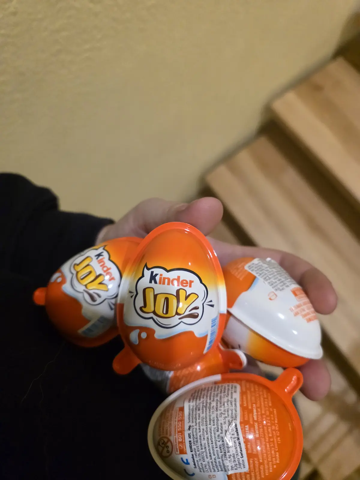 A hand holds multiple Kinder Joy chocolate eggs, some with their orange tops intact and others open, revealing the inner…