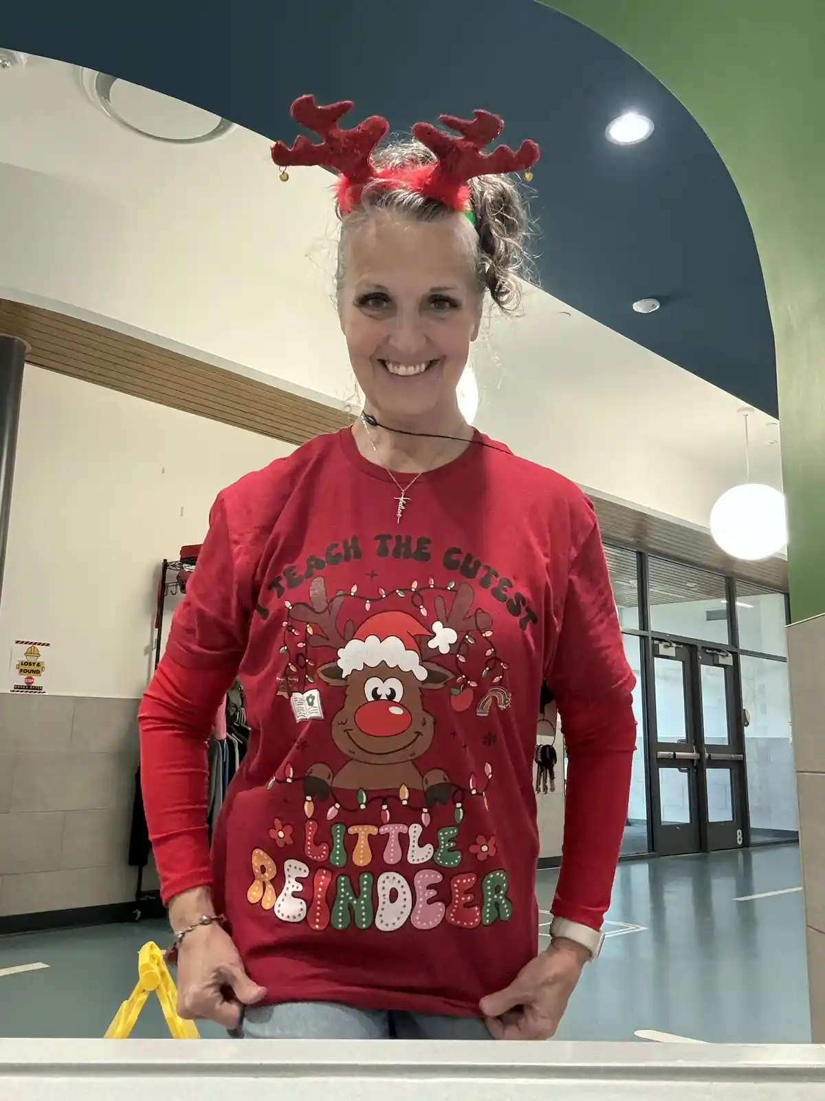 A smiling woman with gray hair in a bun wears red reindeer antlers and a red long-sleeved shirt with a cartoon reindeer…