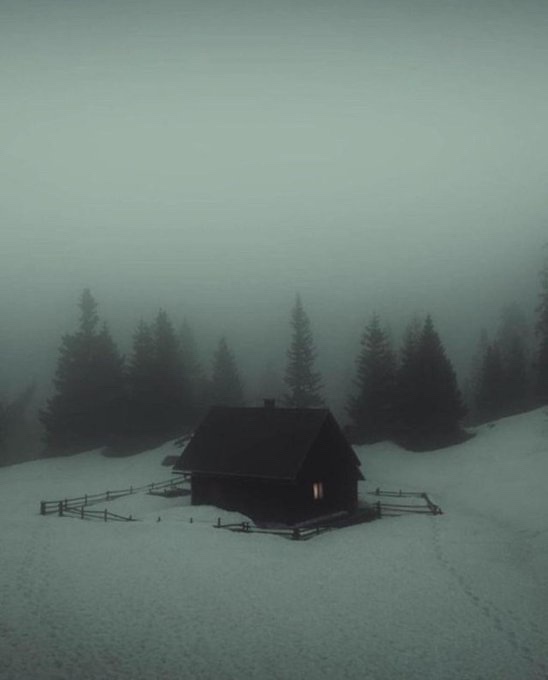 A solitary wooden cabin sits in a snowy, foggy landscape with pine trees in the background. A single warm light glows from a…
