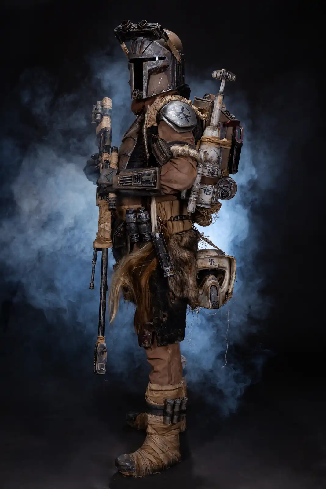 A person in a highly detailed, weathered Mandalorian armor costume stands in a smoky, dark studio setting, facing away from…