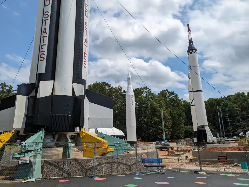 A photo of three large NASA rockets on display outdoors, surrounded by construction fencing and signs. One rocket has…