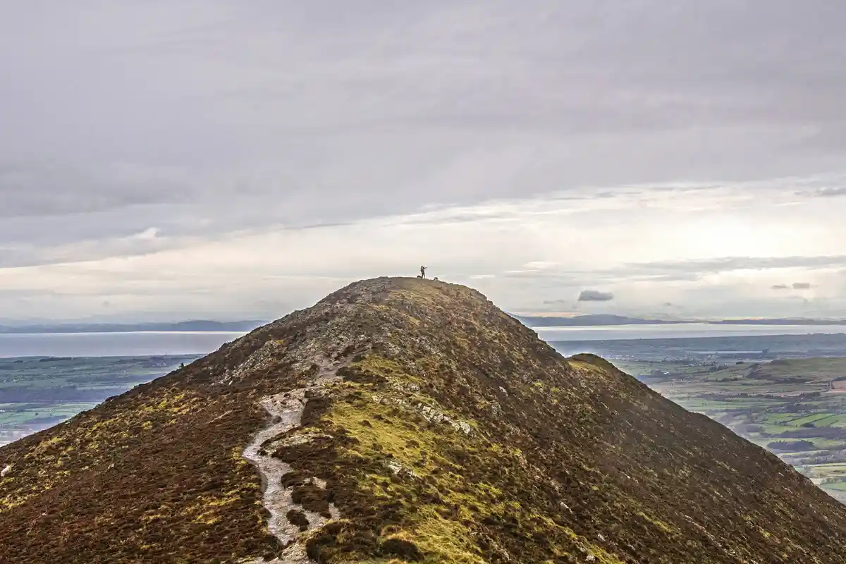 A person stands triumphantly at the peak of a small, grassy hill, arms raised, overlooking a vast landscape with distant…