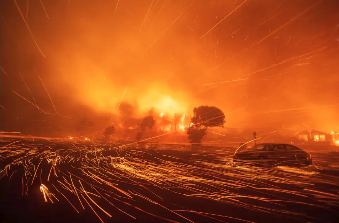 A dramatic photograph shows a car parked in a field during a massive wildfire, with intense orange flames and embers flying…