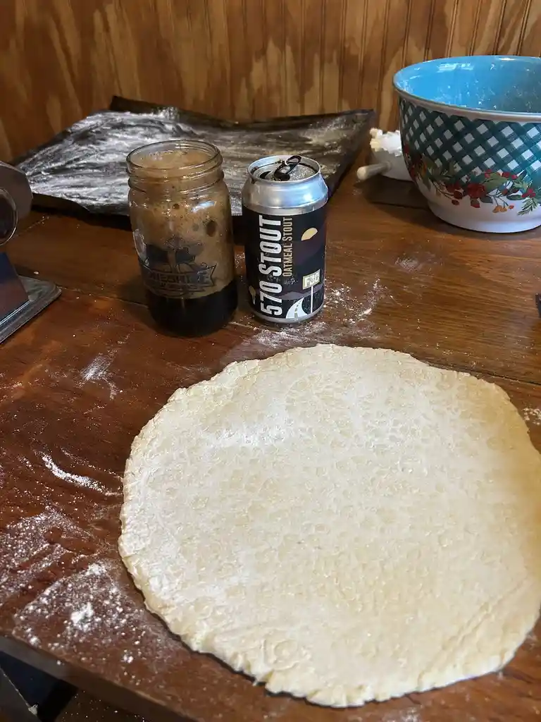 A round, flour-dusted pie crust sits on a wooden table next to an open can of '570 Stout Oatmeal Stout' and a mason jar…