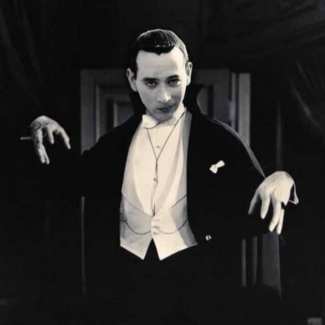 A black-and-white photo of a man in a tuxedo with a cross necklace and claw-like hands, looking menacingly at the camera…