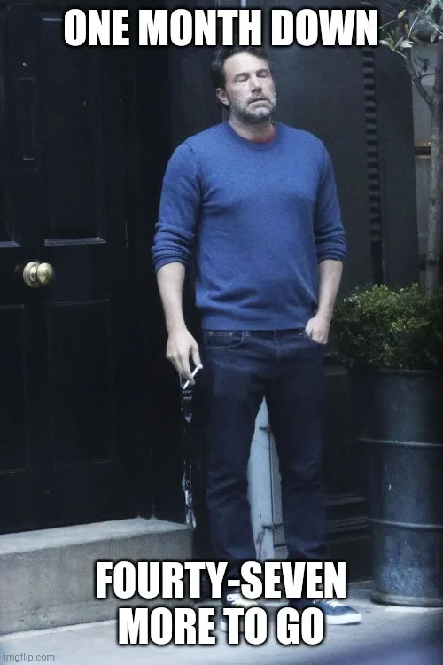 A man in a blue sweater and jeans stands outside a dark door, looking exhausted and defeated while holding a cigarette. The…